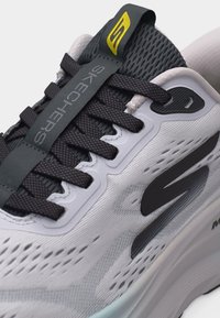 Κοντινή λήψη λευκού και μαύρου αθλητικού παπουτσιού Skechers με μαύρα κορδόνια, γλώσσα από δίχτυ και κίτρινη λεπτομέρεια λογότυπου στην ταμπέλα της γλώσσας.