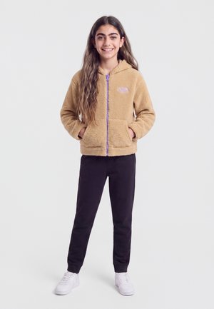 Fille souriante aux longs cheveux, debout les mains dans les poches, portant une veste polaire beige, un pantalon noir et des baskets blanches sur un fond uni.
