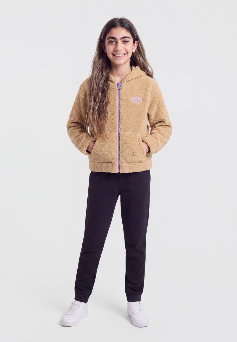 Fille souriante aux longs cheveux, debout les mains dans les poches, portant une veste polaire beige, un pantalon noir et des baskets blanches sur un fond uni.