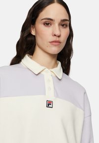 Koszulka polo w białym i jasnofioletowym kolorze, z kołnierzykiem i guzikami, z małym logo marki na piersi. Materiał bawełniany.