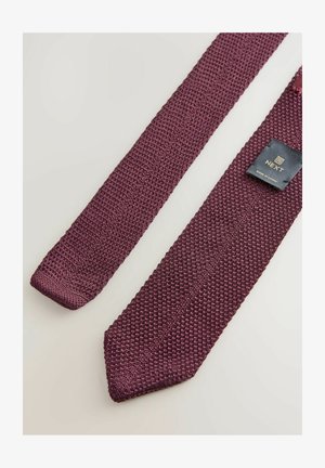 Bordeaux strikket slips med et tekstureret stof, der har et slankt design og et mærke med "NEXT" logo, på en lys baggrund.
