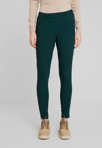 Leggings vert foncé avec une texture lisse, un design ajusté, une taille élastique et des poches latérales, associés à des chaussures beiges.