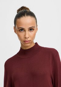 Burgundi magas nyakú pulóver, bordázott textúrával, hosszú ujjak, testhez simuló siluett és minimalista design, látható minták vagy hardver részletek nélkül.