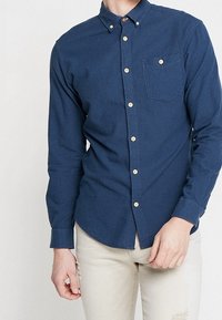 Laivastonsininen napillinen paita, jossa on teksturoitu kuvio, yksittäinen rintatasku ja vaaleat napit, valmistettu kudotusta kankaasta.