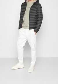 Man i svart vadderad jacka, beige sweatshirt, vita byxor och vita sneakers står mot en enfärgad bakgrund.