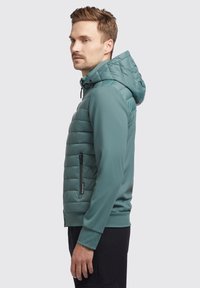 Tarnfarbige gepolsterte Jacke mit taillierter Passform, einer mit Reißverschluss versehenen Tasche auf der linken Seite und einer Kapuze mit verstellbaren Kordeln. Glatte Textur.