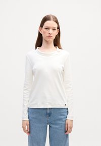 Langarm wit top met een colhals, zachte textuur en een getailleerd ontwerp. Draagt goed met lichtblauwe jeans, met een klein logo aan de zoom.