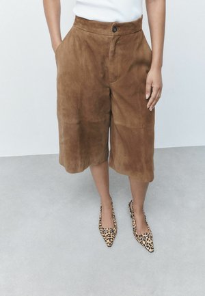 Person iført brune ruskindshorts til knæet, hvid ærmeløs top og spidse leopardmønstrede sko, der står på en grå gulv.