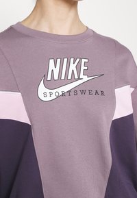 Lila sweatshirt med Nike-logotyp och texten "SPORTSWEAR". Färgblockdesignen inkluderar ljusrosa och mörklila accenter. Mjuk tygkänsla.