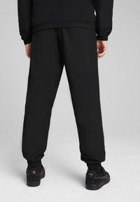 Pantalons de sport noirs avec une taille élastique et des poignets, présentant une texture lisse. Accent rouge notable sur le côté.