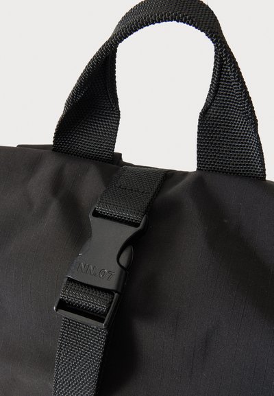 Sac en tissu noir avec poignée tissée et sangle réglable équipée d'une boucle en plastique noire portant la mention NN.07 sur un fond neutre.