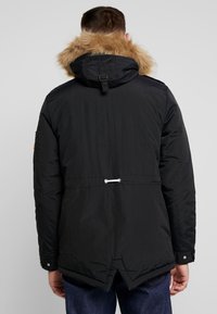 Svart parkas med foder av fuskpäls, med indragen midja, dragsko-detaljer och tryckknappar på ärmarna. Texturerad tyg.