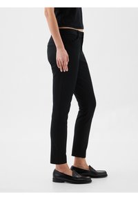 Personne portant un pantalon noir slim-fit longueur cheville et des mocassins noirs, debout de profil sur un fond uni.