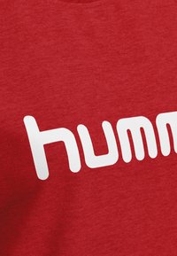 T-shirt in cotone rosso con un grande logo bianco in lettere arrotondate che scrive "humm." Il tessuto appare liscio con una trama media.