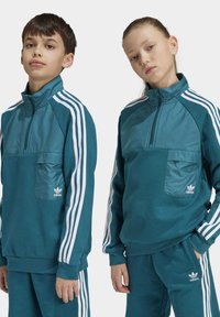Dos niños de pie uno al lado del otro, vistiendo chándales Adidas a juego en color teal con rayas blancas y camisetas de media cremallera con bolsillos frontales.