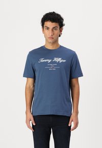 Sötétkék pamut póló rövid ujjakkal. Fehér nyomtatott logó és a "Tommy Hilfiger ESTABLISHED 1985 NEW YORK" felirat található rajta. Kerek nyakkivágású.