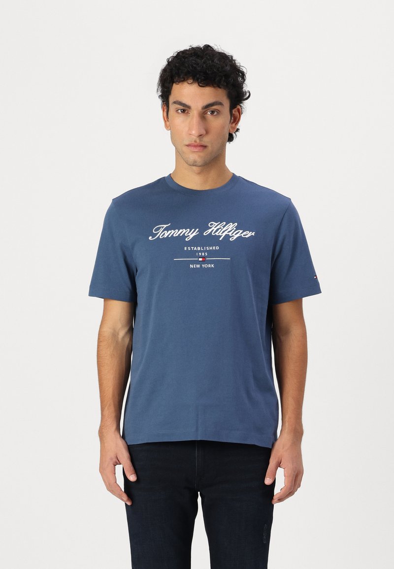 Sötétkék pamut póló rövid ujjakkal. Fehér nyomtatott logó és a "Tommy Hilfiger ESTABLISHED 1985 NEW YORK" felirat található rajta. Kerek nyakkivágású.