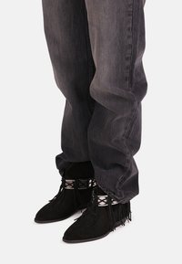 Črne ankel čevlje iz nubuk usnja z ostrimi prsti in detajli z fru fruji, v kombinaciji s sivo jeans hlačami. Čevlji imajo dekorativno vezanje in srebrne poudarke.