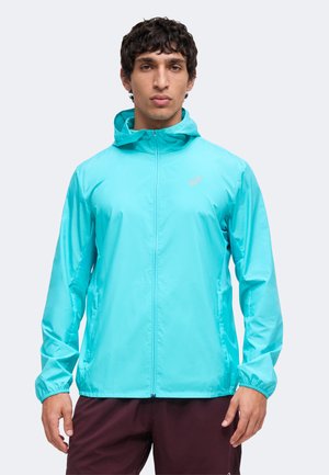 Homme portant une veste coupe-vent à capuche turquoise et un short foncé, debout devant un fond clair uni.