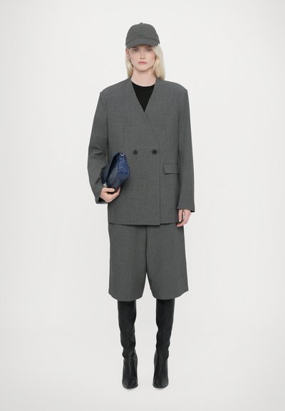 Blazer grigio su misura a doppio petto su una maglia nera, abbinato a pantaloni corti grigi e stivali neri alti fino al ginocchio. Tiene una clutch strutturata di colore blu scuro.