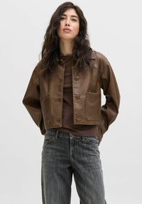 Giacca cropped in pelle marrone con due tasche frontali, chiusura a bottone e una texture liscia, indossata sopra un top marrone e jeans grigi.