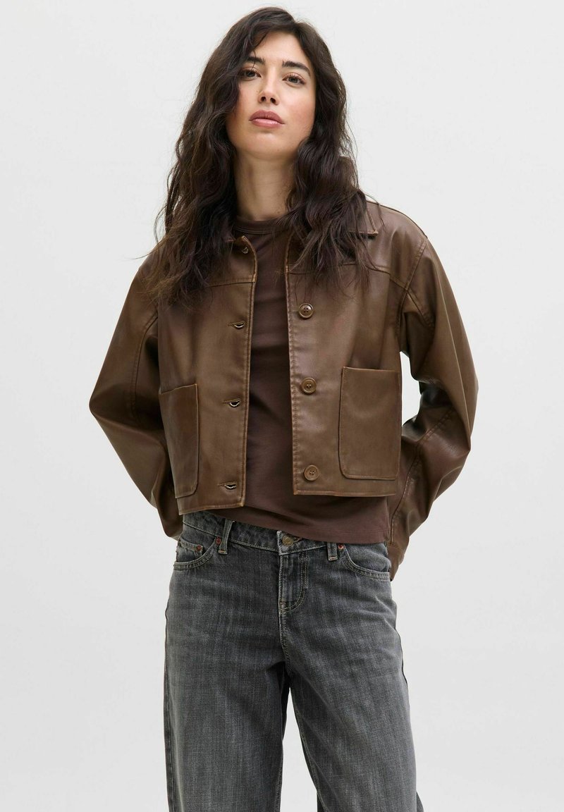 Giacca cropped in pelle marrone con due tasche frontali, chiusura a bottone e una texture liscia, indossata sopra un top marrone e jeans grigi.