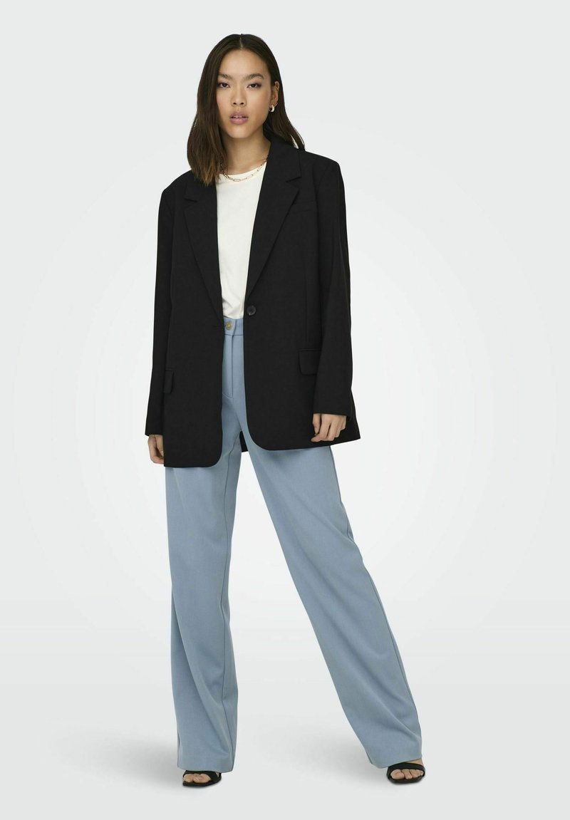 Übergroßer schwarzer Blazer mit zwei Vorderseiten-Taschen, kombiniert mit hellblauen weiten Hosen. Darunter ein schlichtes weißes T-Shirt und schwarze High Heels.