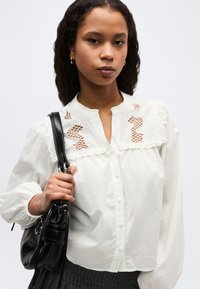 Νεαρή γυναίκα με λευκή μπλούζα με δαντελένιες λεπτομέρειες στο γιακά, κρατώντας μια μαύρη δερμάτινη τσάντα ώμου.