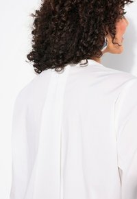 Blouse blanche avec un design simple et minimaliste. Dispose d'une couture centrale au dos et d'une texture douce et lisse.