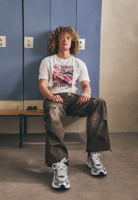 Valge graafiline T-särk punase ja musta printimisega, pruunid cargo-püksid ja hõbedased sneakerid mustade aktsentidega. Kiharaid juukseid, istub puidust pingil.