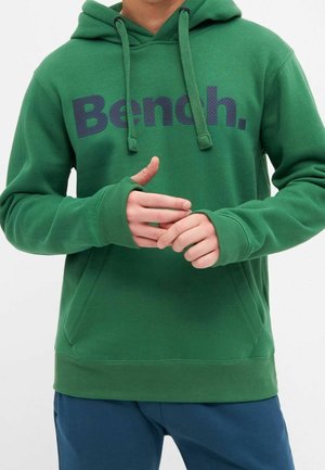 Zielona bluza z kapturem z logo „Bench.” w ciemnych literach, z sznurkami i otworami na kciuki, noszona przez osobę poprawiającą ręce.