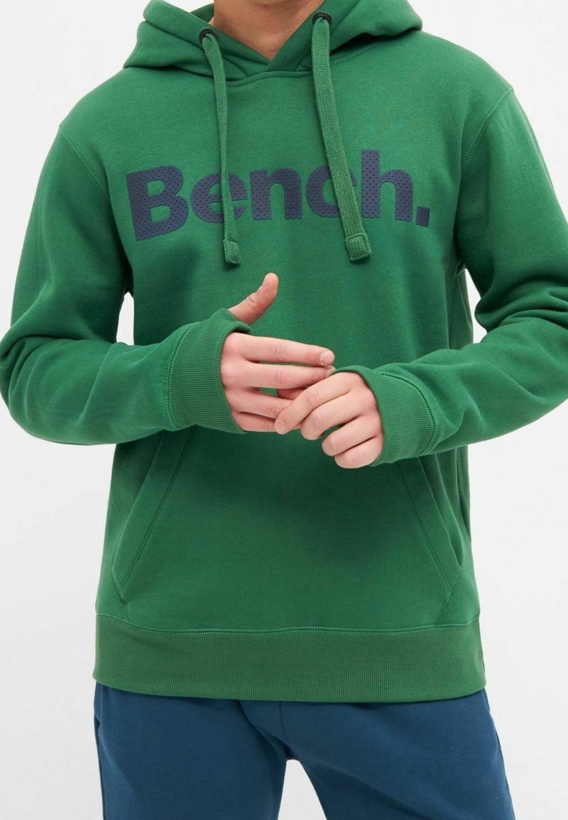 Sweat à capuche vert avec le logo « Bench. » en lettres foncées, avec cordons et trous pour les pouces, porté par une personne ajustant ses mains.