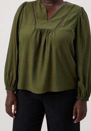 Vrouw draagt een olijfgroene blouse met lange mouwen met geplooide details en een zwarte broek, staand tegen een effen achtergrond.