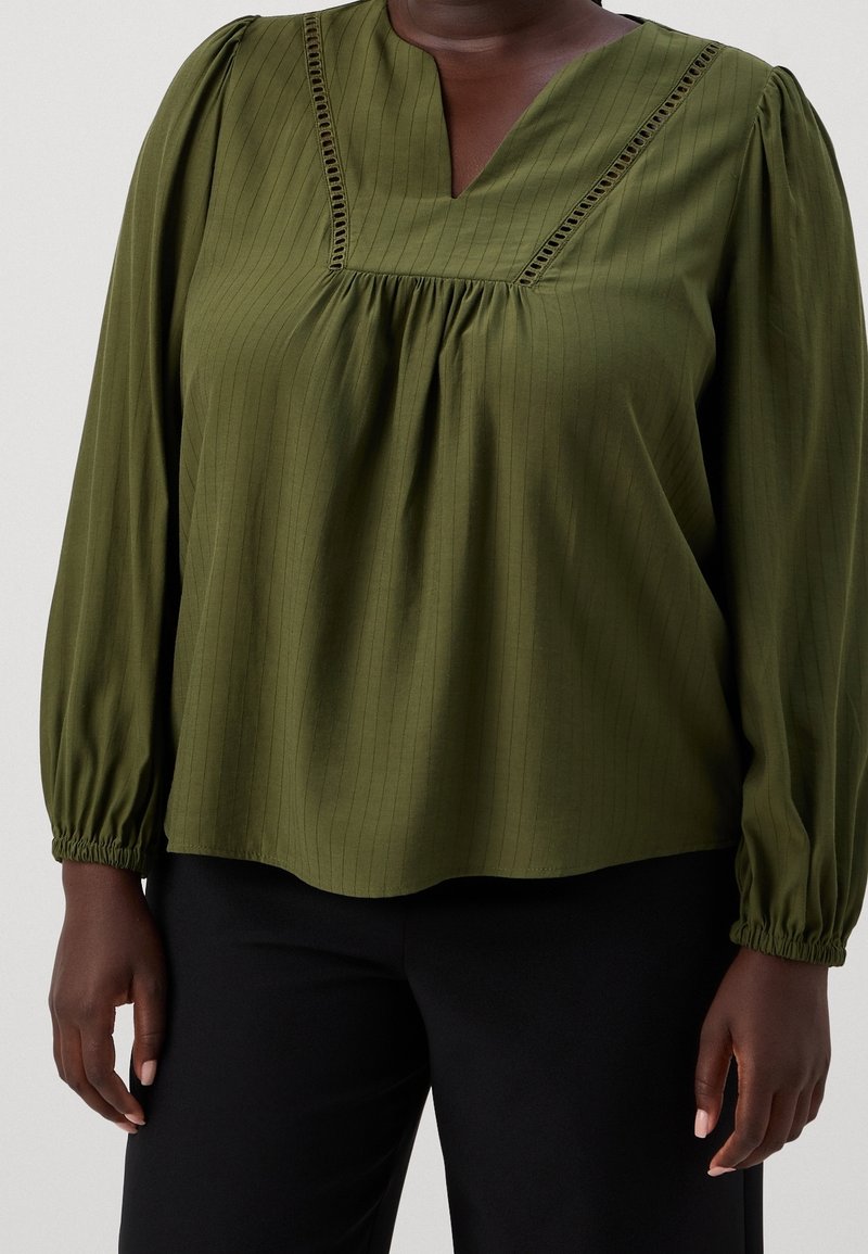 Femme portant un chemisier vert olive à manches longues avec des détails froncés et un pantalon noir, debout devant un fond uni.