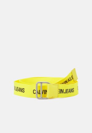 Jaskrawożółty materiałowy pasek z czarnym napisem "Calvin Klein Jeans" i prostokątną, srebrną metalową klamrą, zwinięty na białym tle.
