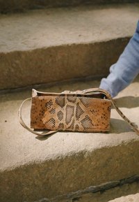 Python leren handtas in bruintinten met een reptielenpatroon en een dunne schouderriem, rustend op een betonnen oppervlak met zichtbare textuur.