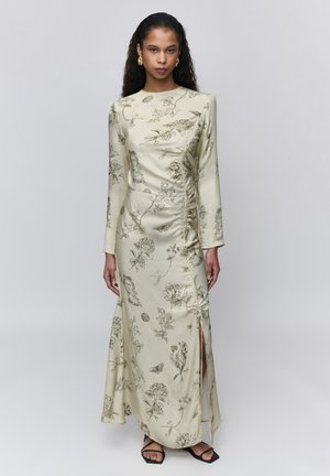 Kobieta ubrana w długą kremową sukienkę z czarnymi kwiatowymi szkicami, marszczonymi detalami po bokach, rozcięciem do uda, złotymi kolczykami kołami i czarnymi sandałami.