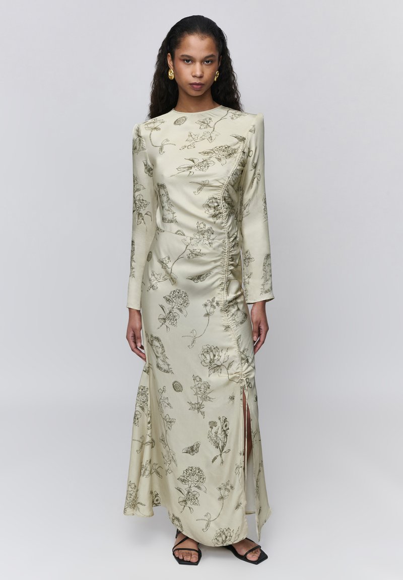 Femme portant une longue robe crème avec des croquis floraux noirs, un détail froncé sur le côté, une fente haute sur la cuisse, des boucles d'oreilles anneaux dorées et des sandales noires.