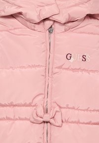 Rožā kvadrātu jaka ar rāvējslēdzēju, mazs loks pie jostas, un izšūts "GUESS" logo ar sirds akcentiem uz krūts.