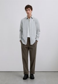 Jasnoniebieska i biała koszula w kratę na guziki, noszona na białym t-shircie, zestawiona z brązowymi spodniami i czarnymi butami, stojąca na neutralnym tle.