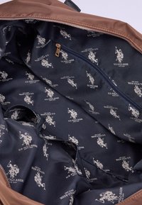 Hnědá látková taška přes rameno s námořnickým podšívkou, která obsahuje vzor bílého loga U.S. Polo Assn. a kapsu na zip. Měkká struktura.