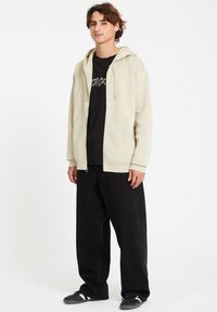 Beige Kapuzenjacke mit Reißverschluss und Kordelzügen, getragen über einem schwarzen T-Shirt. Locker sitzende schwarze Hosen und schwarze Sneakers runden das Outfit ab.