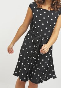 Mustat pilkulliset mekko, jossa on istuva yläosa, lyhyet hihat ja sidottu vyötärö. Laaja helma ja kevyet kangas, jolla on sileä rakenne.