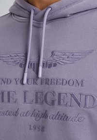 Lilac hoodie featuring an embroidered design with wings and text. Includes drawstrings with plastic aglets and a textured fabric.