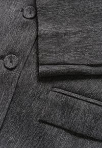 Grau gestepptes Stoffmuster im Fischgrätdesign mit zwei runden Knöpfen, hochgeschlagenen Ärmeln und einem einzelnen Taschendetail im Blazer-Design.