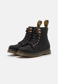 Pár černých kožených šněrovacích bot s bronzovými očky, žlutým prošíváním na podrážce a označenými poutky na zadní straně na bílém pozadí.