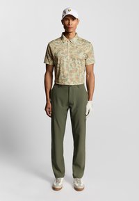 Kortärmad polotröja med ett ljusgrönt blommönster, i kombination med olivgröna byxor och vita golfsandaler, kompletterad med en vit keps.