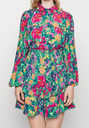 Frau trägt ein langärmeliges, mit Kragen versehenes, geblümtes Kleid mit leuchtend pinken, grünen und gelben Blumen sowie eine taillierte, geraffte Taille und einen Rock.