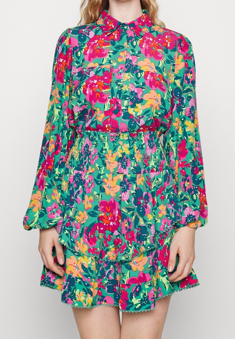 Femme portant une robe fleurie à manches longues et col, avec des fleurs roses, vertes et jaunes vives, et une taille et une jupe cintrées et à volants.