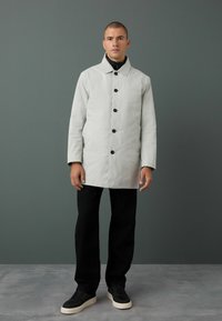 Abrigo de color gris claro, con botones y un collar estructurado, superficie texturizada y botones negros, combinado con pantalones negros y zapatillas.